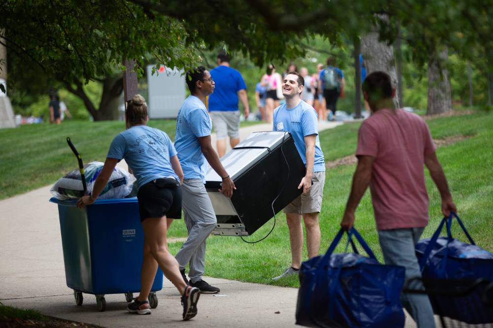 Two alumni carrying a mini fridge into a student's dorm.